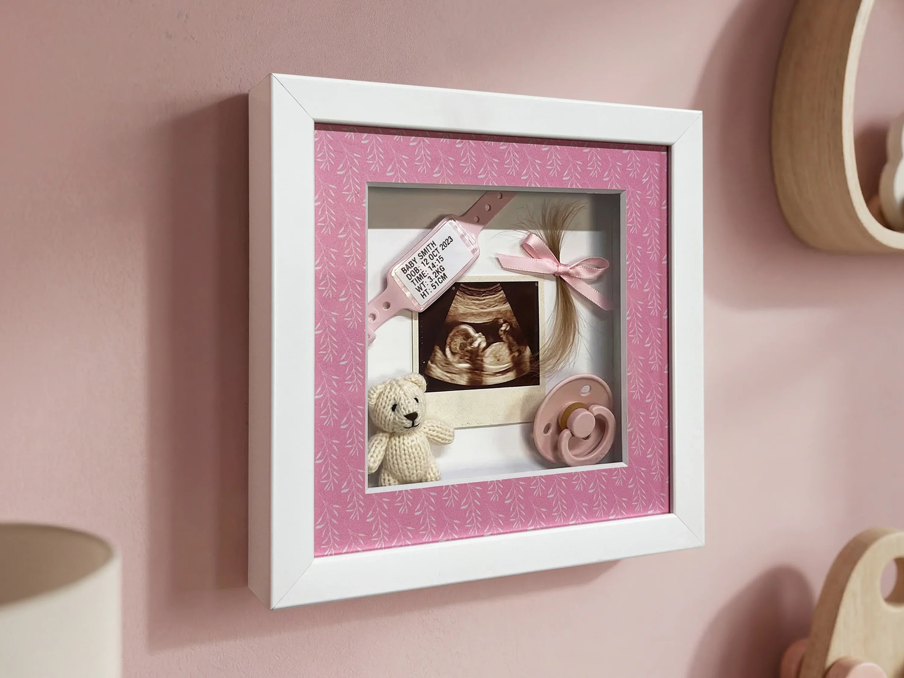 Pink and white shadow box frame displaying baby keepsakes, ultrasound, pacifier, and teddy bear